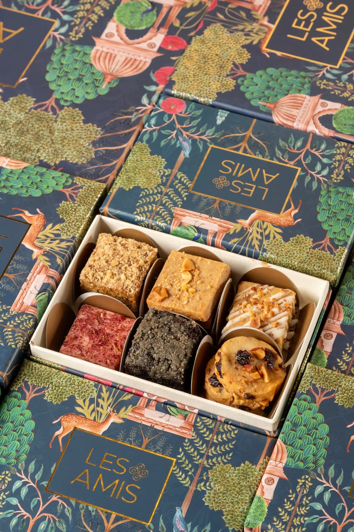 An open tray of six Les Amis Diwali sweets — pedas, kalakand and tea cakes — on the navy illustrated hamper boxes